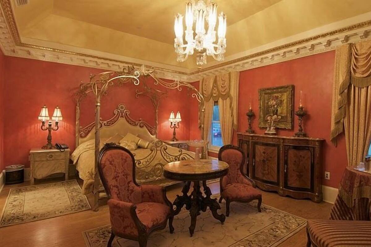 A room with a four-poster bed, two armchairs, a round table, and a chandelier.