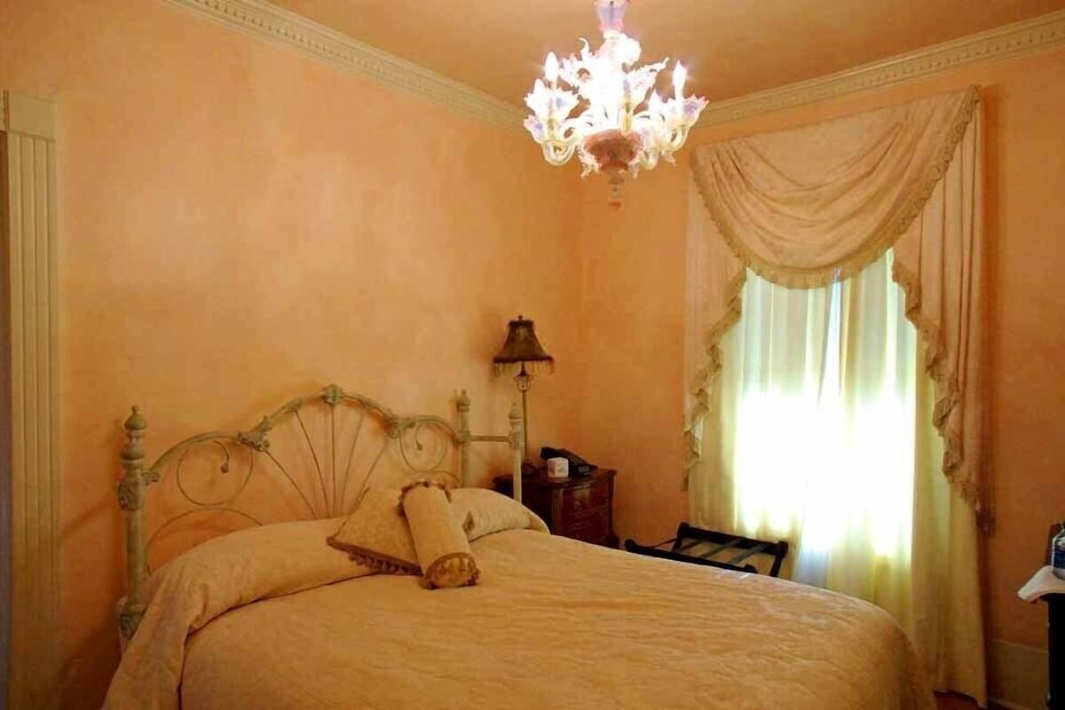 A bedroom with a bed, a chandelier, a nightstand, and a window with curtains.