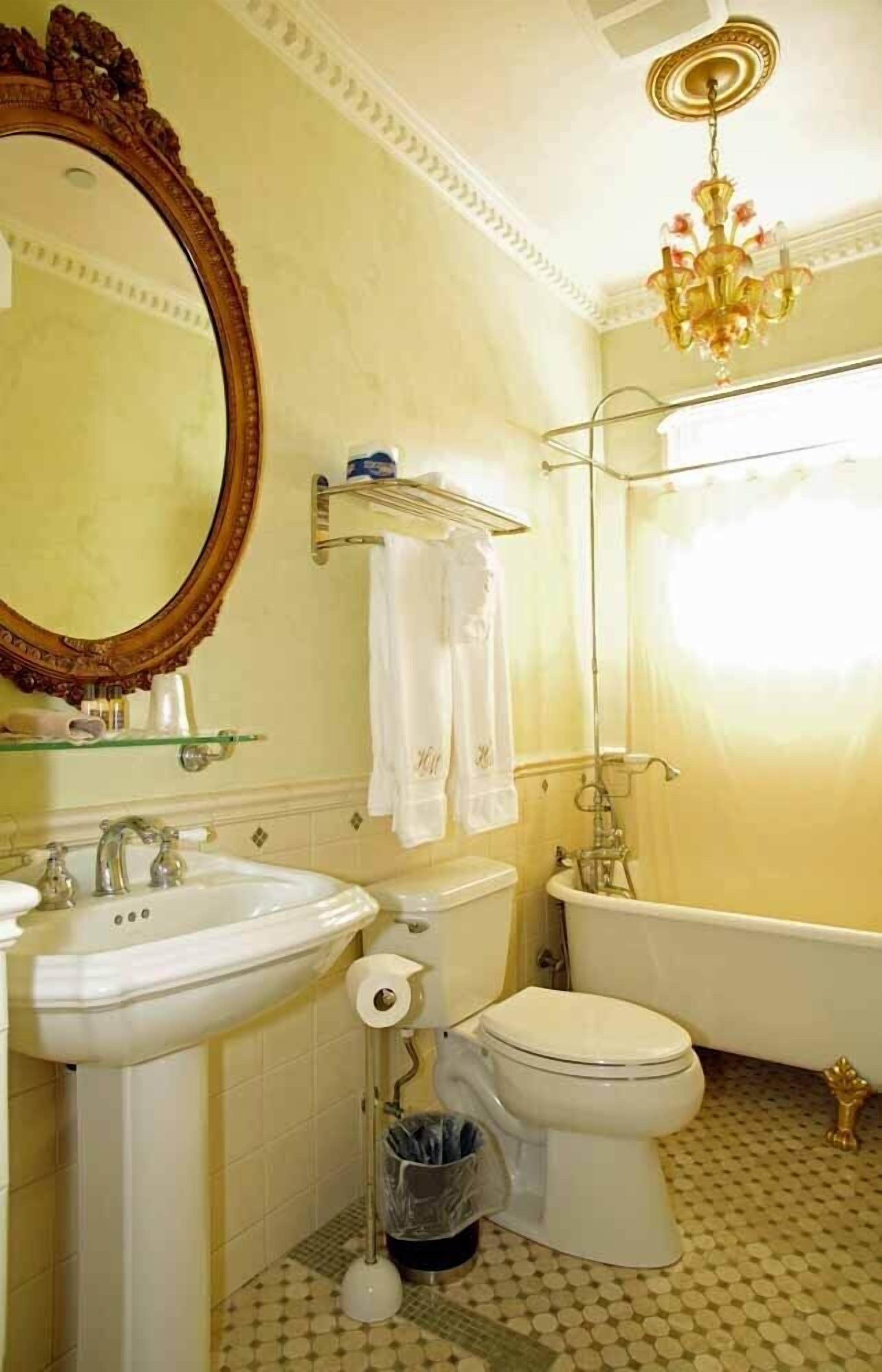 Ein Badezimmer mit Waschbecken, Toilette und Badewanne. Über dem Waschbecken hängt ein Spiegel, über der Badewanne eine Chandelüster.