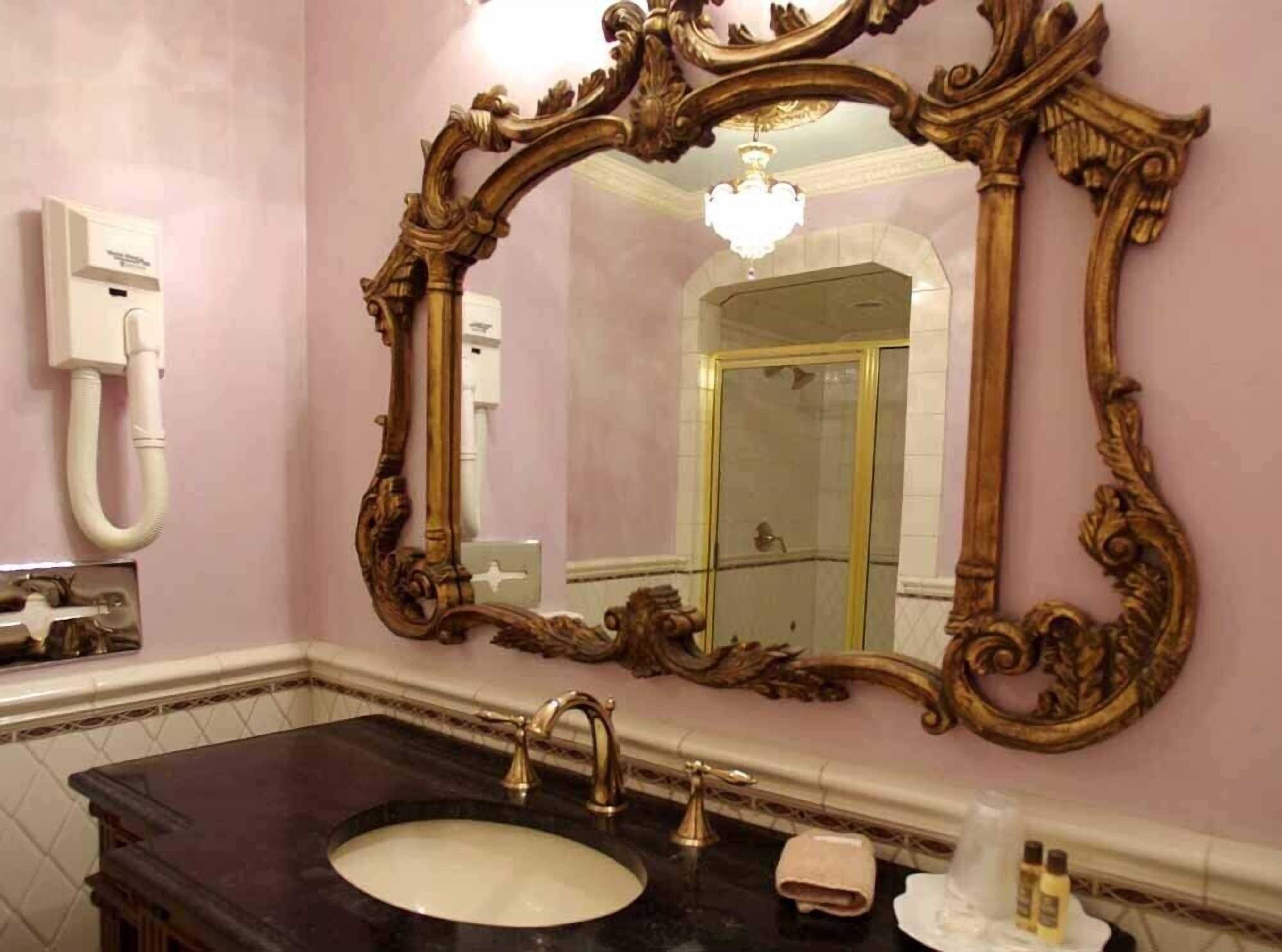 Ein Badezimmer mit einem großen, verzierten Spiegel, einem schwarzen Waschbecken und einer sichtbaren Dusche im Hintergrund.
