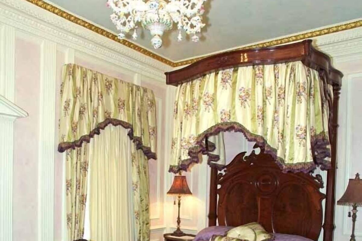 A bedroom with a four-poster bed, two upholstered chairs, a chandelier, and a patterned rug.