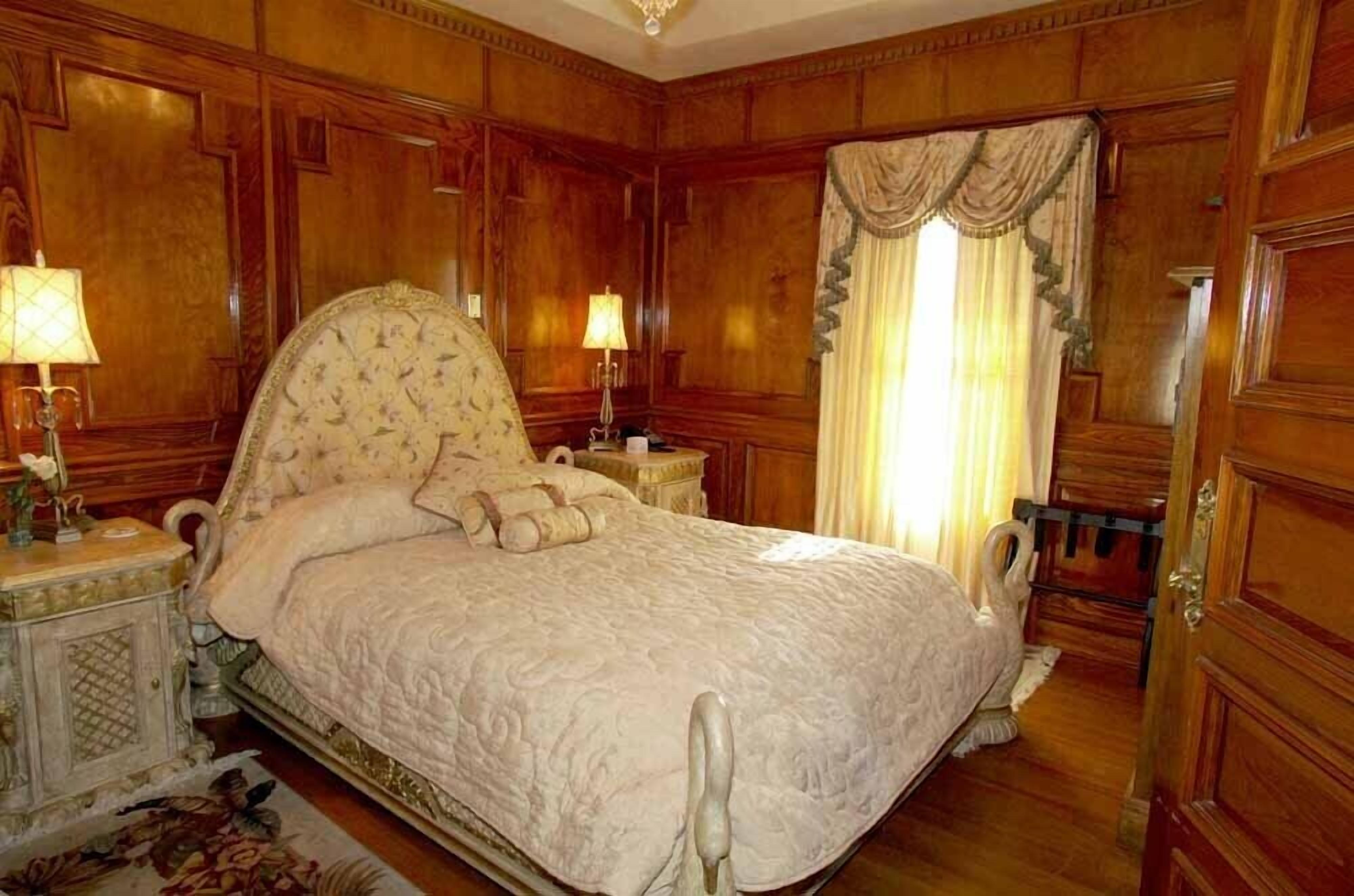 Un letto a baldacchino classico in legno con copripiumino bianco, due comodini con lampade e un'ampia finestra con tende.