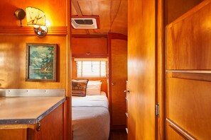 A compact, wood-paneled interior with a bed, a small table, a painting, and a window with blinds. - The Vintages Trailer Resort (Dayton)