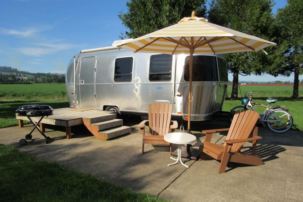A silver RV parked on a concrete patio with a picnic table, two Adirondack chairs, and a striped umbrella.