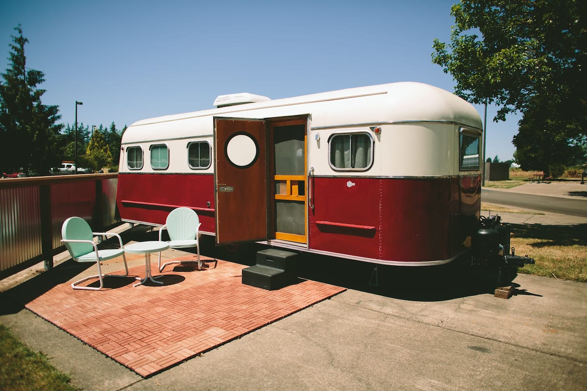 A stationary RV with a door open, revealing a small seating area with two chairs and a table, and a step leading to the entrance.