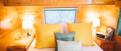A neatly made bed with yellow and patterned pillows, a bedside table with a lamp and a radio, and wooden headboard and walls.