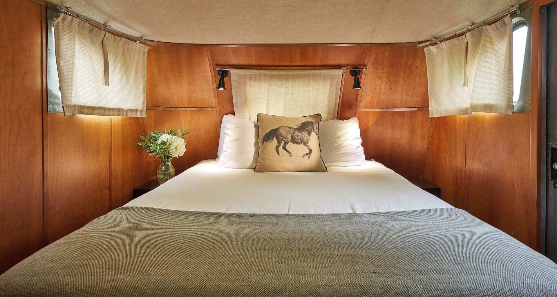 Un dormitorio con cabecera de madera, cama con ropa de cama blanca, almohada con tema de caballos y cortinas.