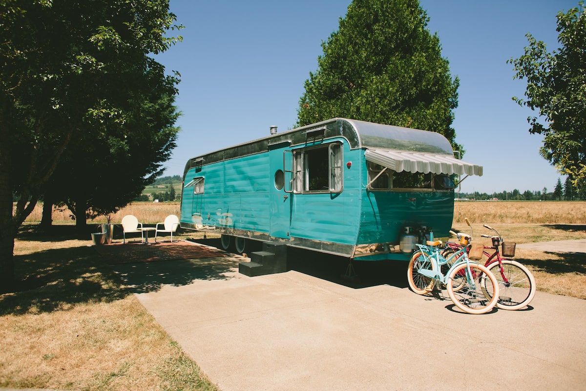A turquoise trailer with a covered front, two bicycles, and a picnic table in a grassy area.