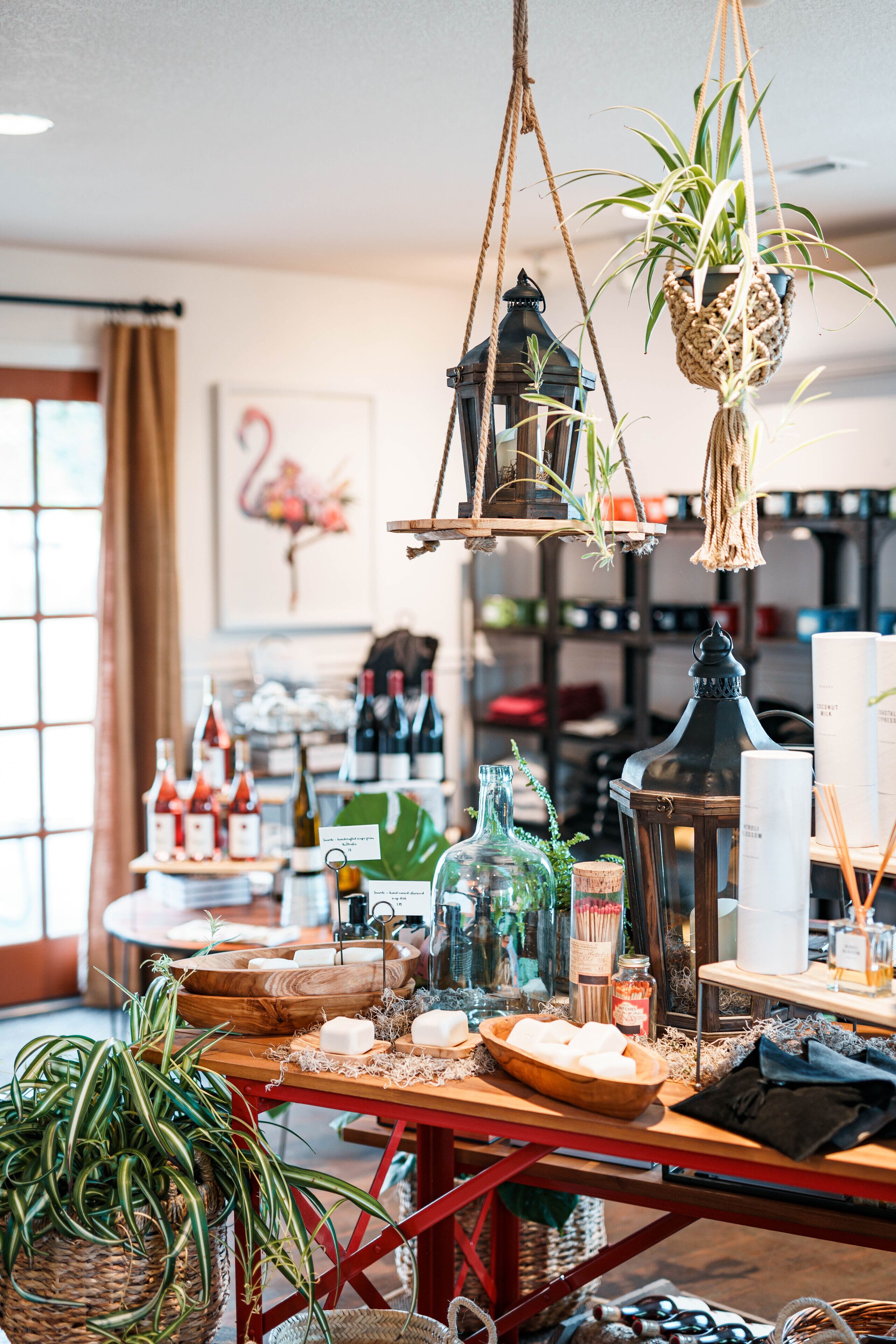 A room with a wooden table displaying various bottles and candles, a hanging plant, and a bookshelf in the background.