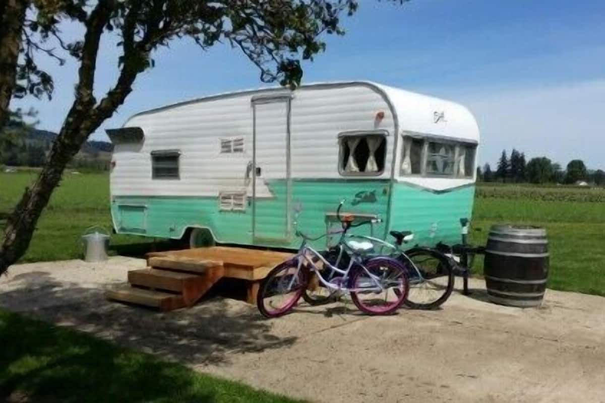 A vintage camper with a bicycle parked in front, a picnic table, and a barrel.