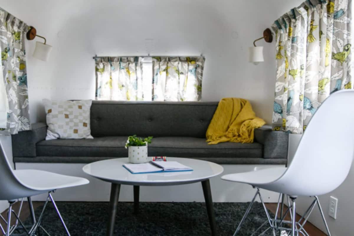 A cozy living area with a grey sofa, two white chairs, a small round table with a notebook and pen, and a yellow throw blanket.