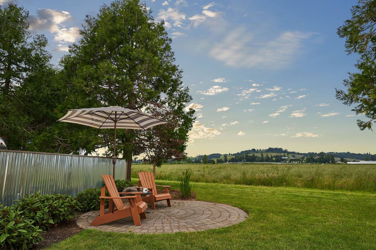 An outdoor patio with two wooden chairs and a striped umbrella, surrounded by a metal fence and lush greenery.
