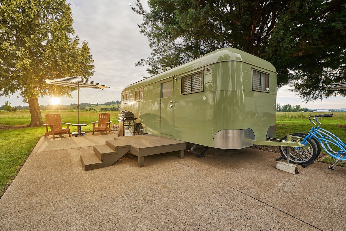 A green RV with a patio area, including wooden chairs, a table, and a bicycle.