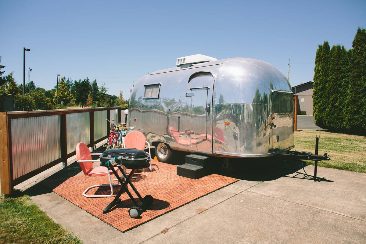 A silver trailer with a small window and door, parked on a brick patio with a grill and chairs.