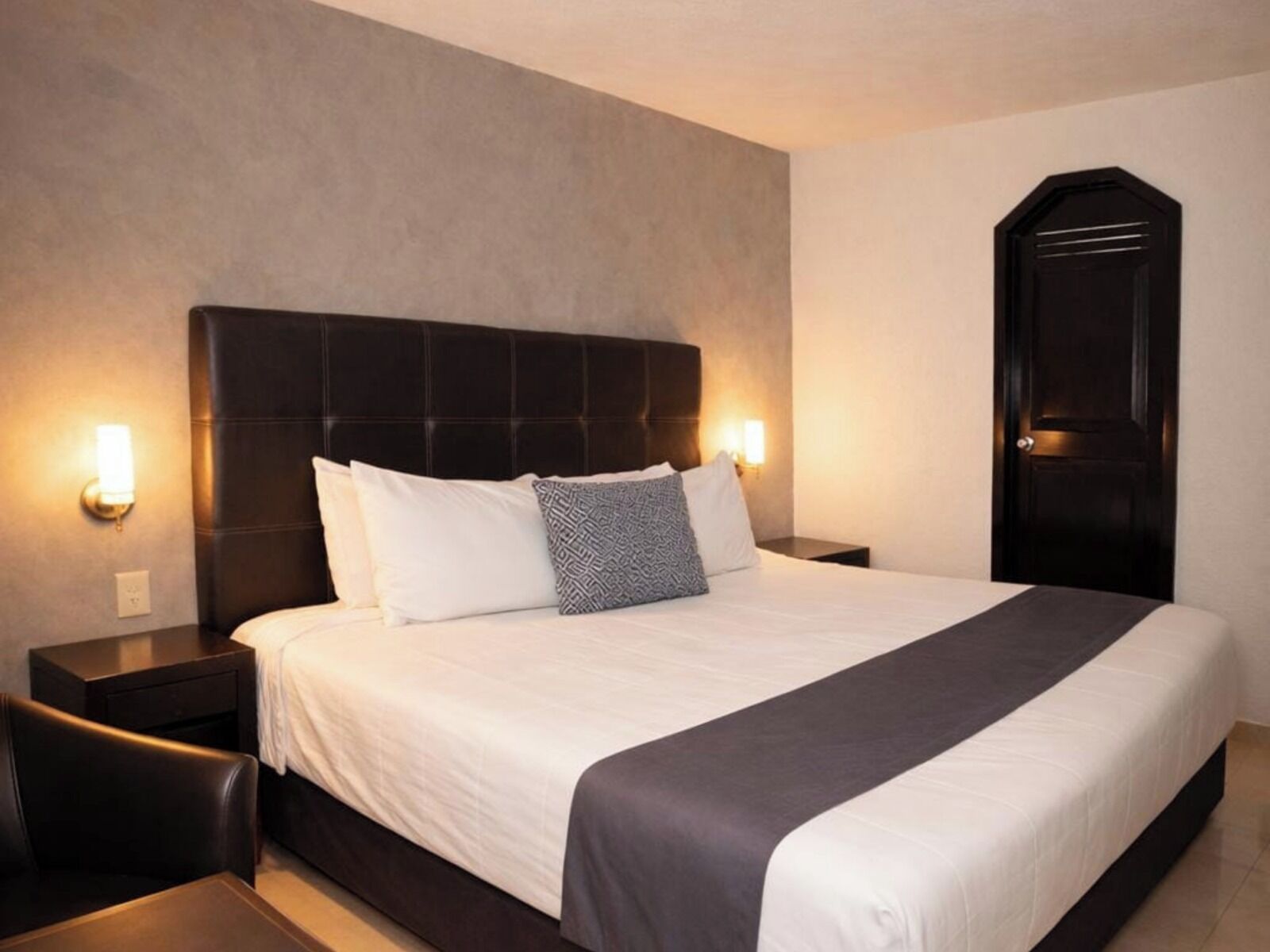 Una habitación de hotel con una cama grande, dos mesitas de noche, un cabecero oscuro y una puerta.