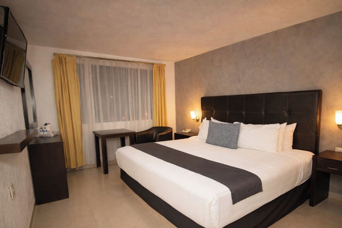 A hotel room with a large bed, bedside tables, a desk, a chair, and a television.