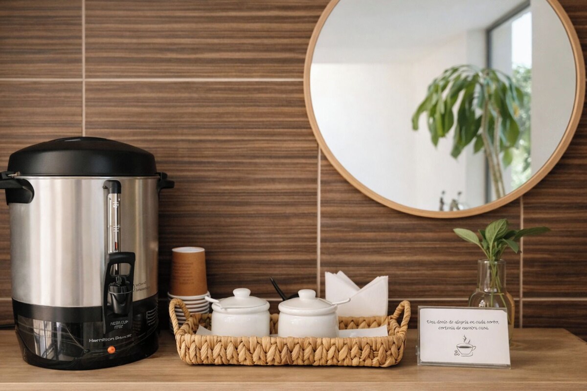 A kitchen area with a stainless steel electric kettle, a woven basket with white containers, a mirror, and a sign with a coffee cup illustration.