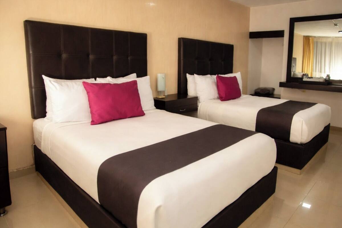 Two double beds with black headboards and white bedding, a nightstand with a lamp, and a large mirror above the beds.