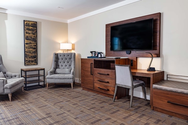 Une chambre d’hôtel dotée d’un grand téléviseur à écran plat, d’un fauteuil à capitons gris, d’un bureau en bois avec des tiroirs et d’un tapis à motifs.