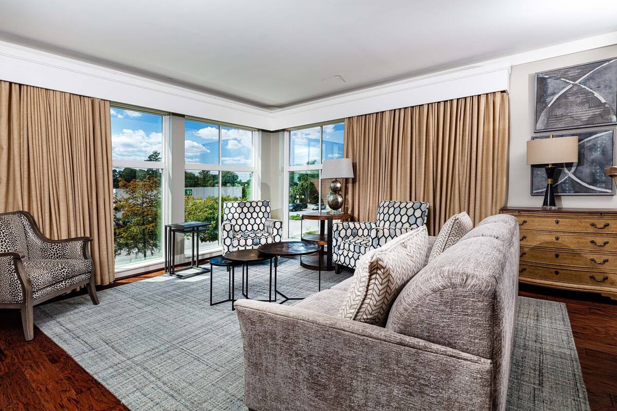 Un amplio salón con sofá, sillones, una mesa baja y una mesita auxiliar. Grandes ventanales ofrecen vistas al exterior.