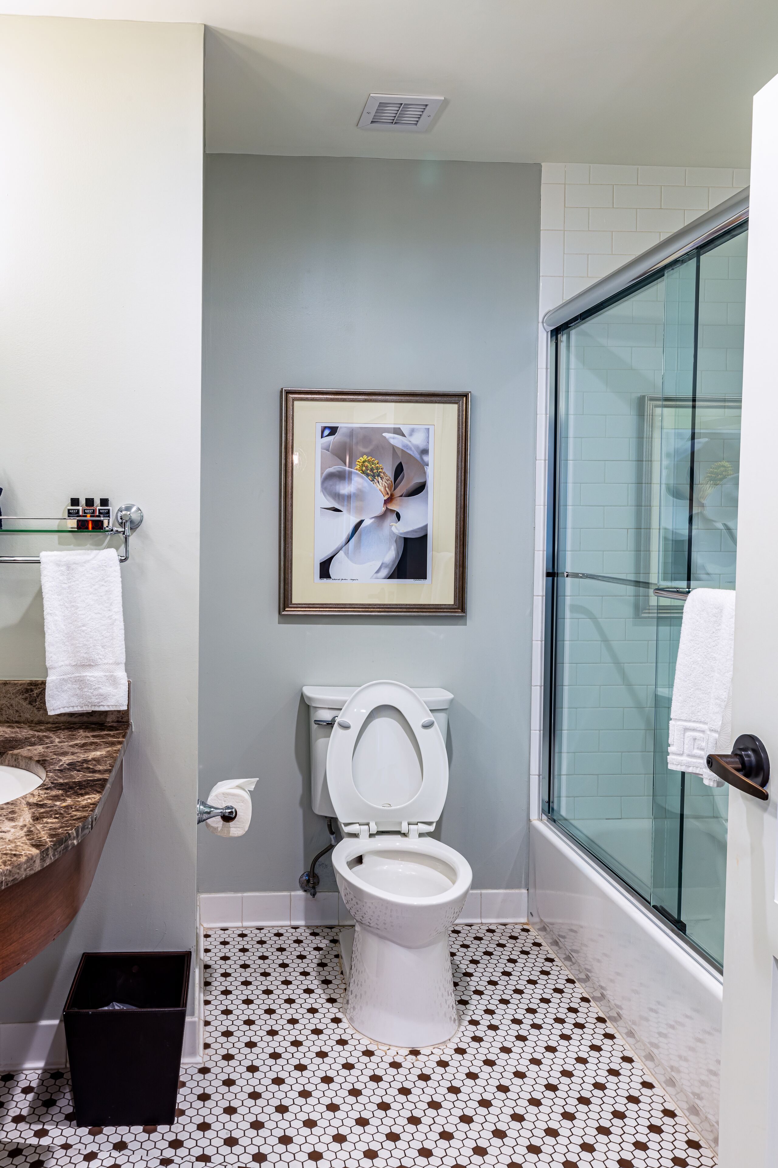 A bathroom with a toilet, a framed picture on the wall, a glass shower enclosure, and a tiled floor.