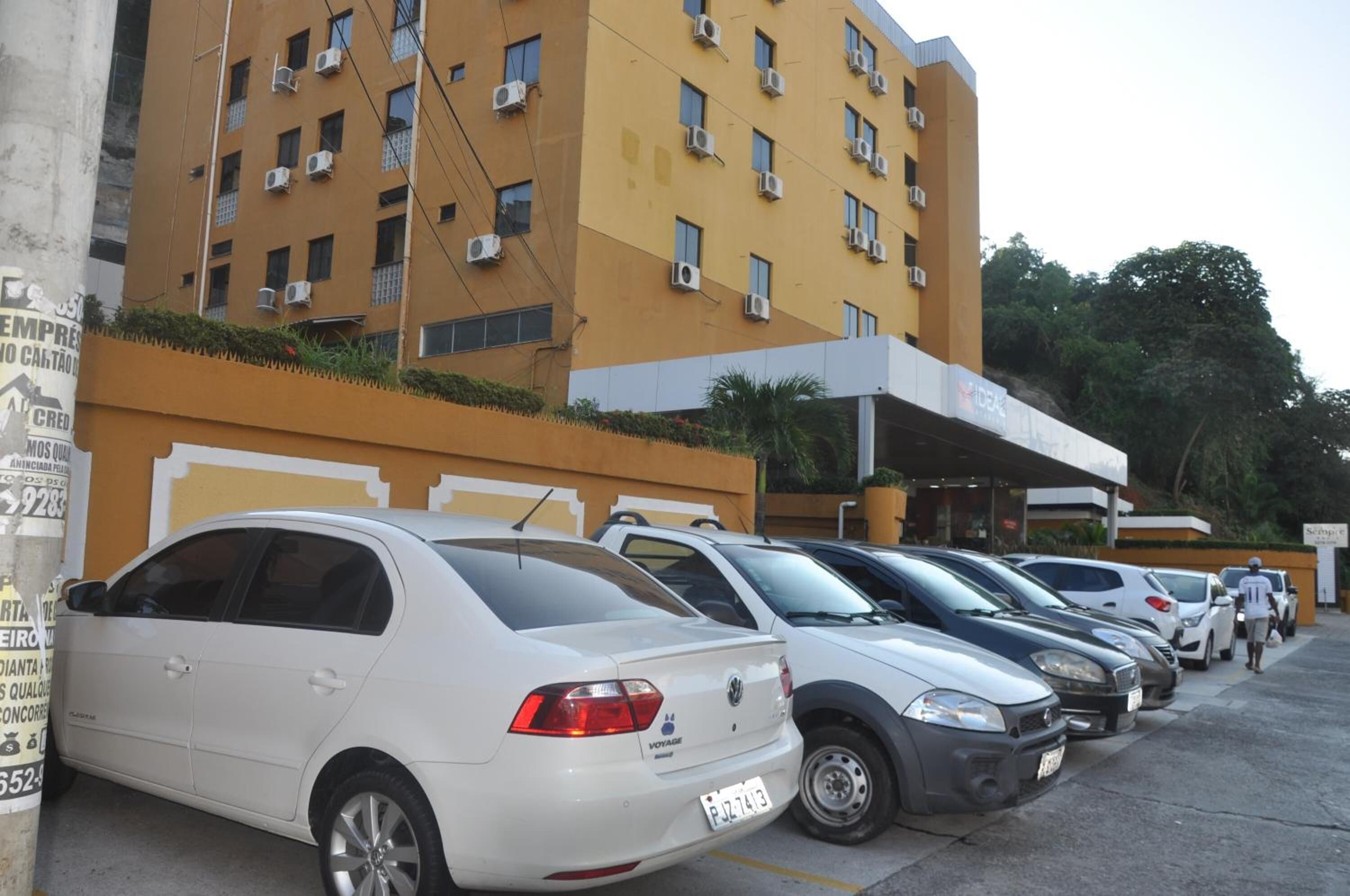 A row of parked cars in front of a yellow building with multiple air conditioning units.