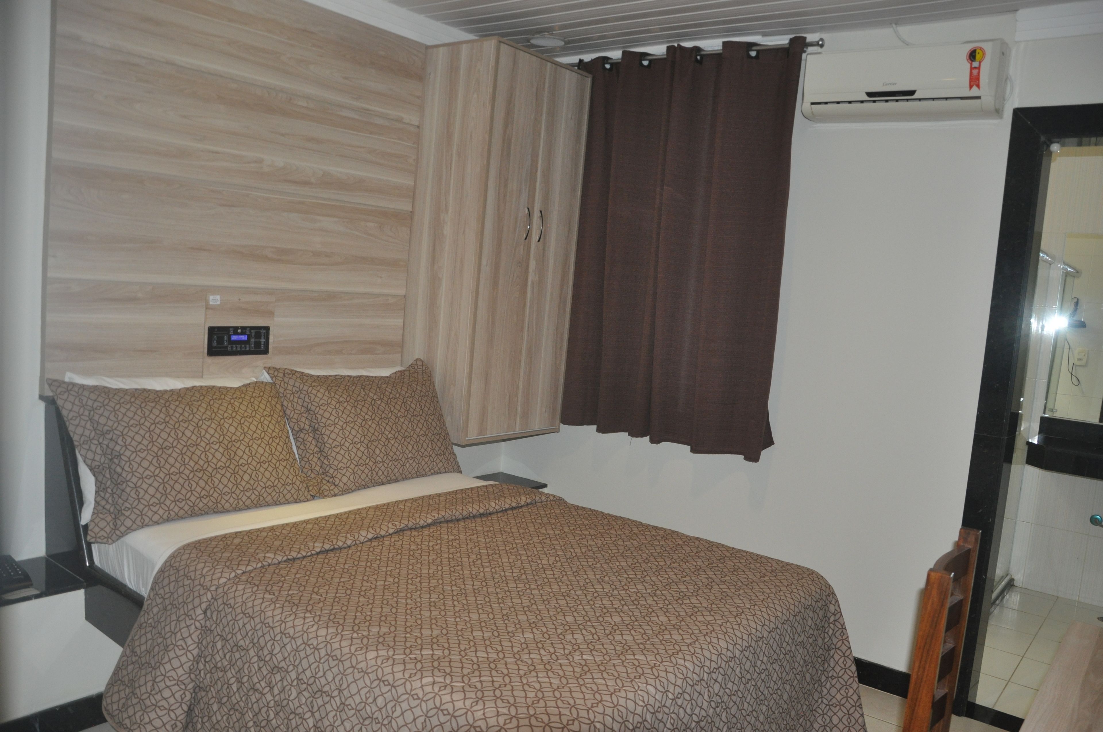 Un dormitorio con cama, mesita de noche, reloj digital, ventana con cortinas y un equipo de aire acondicionado.