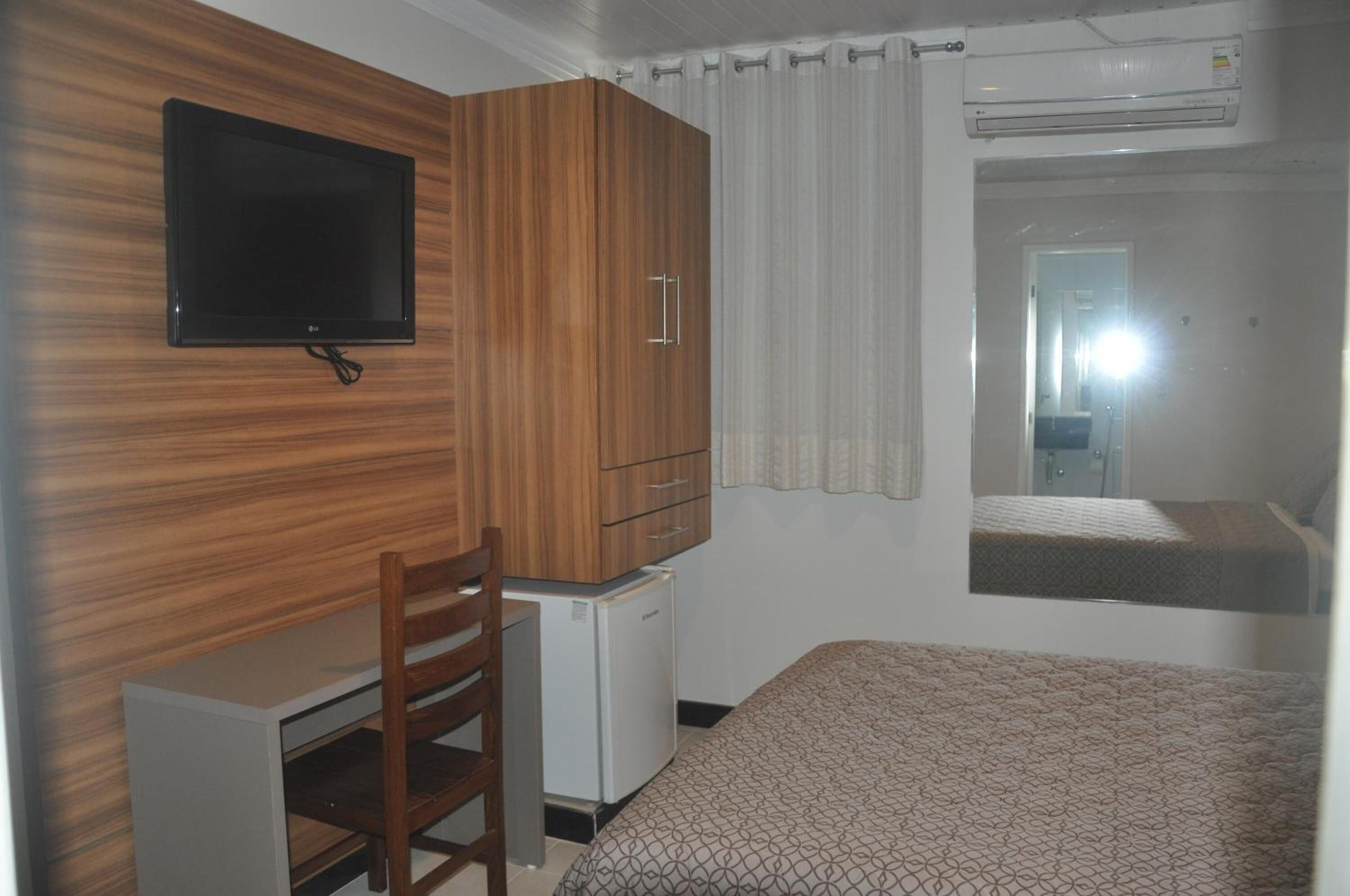 Un dormitorio con una cama, un escritorio con silla, un televisor montado en la pared y un armario.