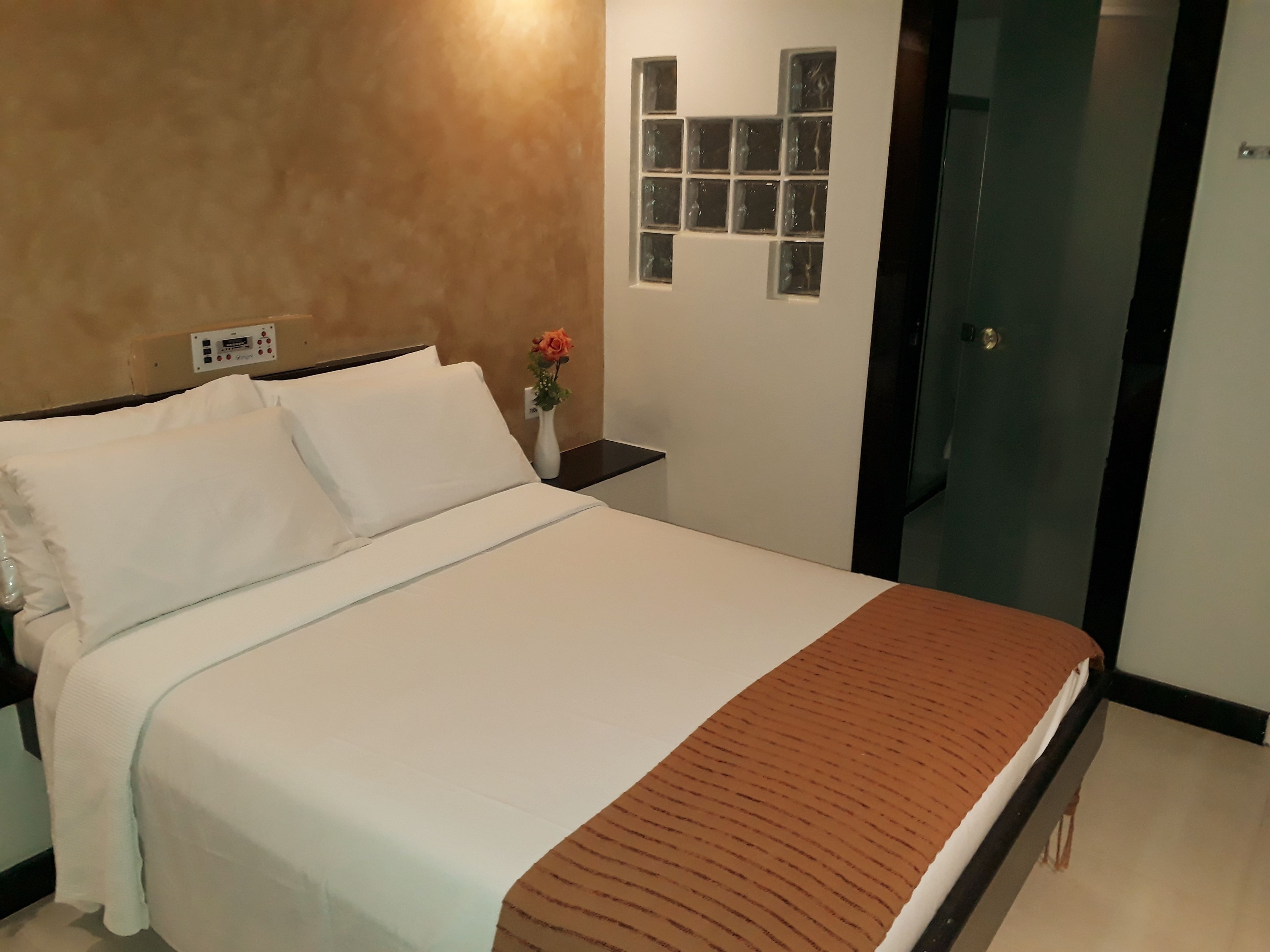 A neatly made bed with white linens and a brown blanket, a bedside table with a vase of flowers, and a wall-mounted light switch.