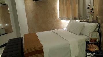 A hotel room with a bed, a bedside table with a phone and a menu, a bench, an air conditioning unit, and a window with curtains.