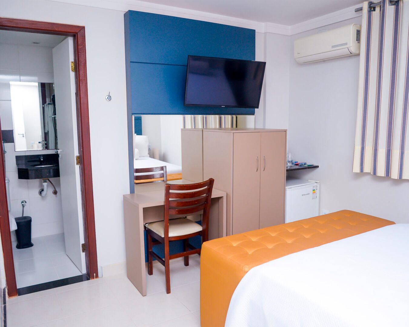 Habitación de hotel con cama, escritorio con silla, televisor, armario y aire acondicionado.