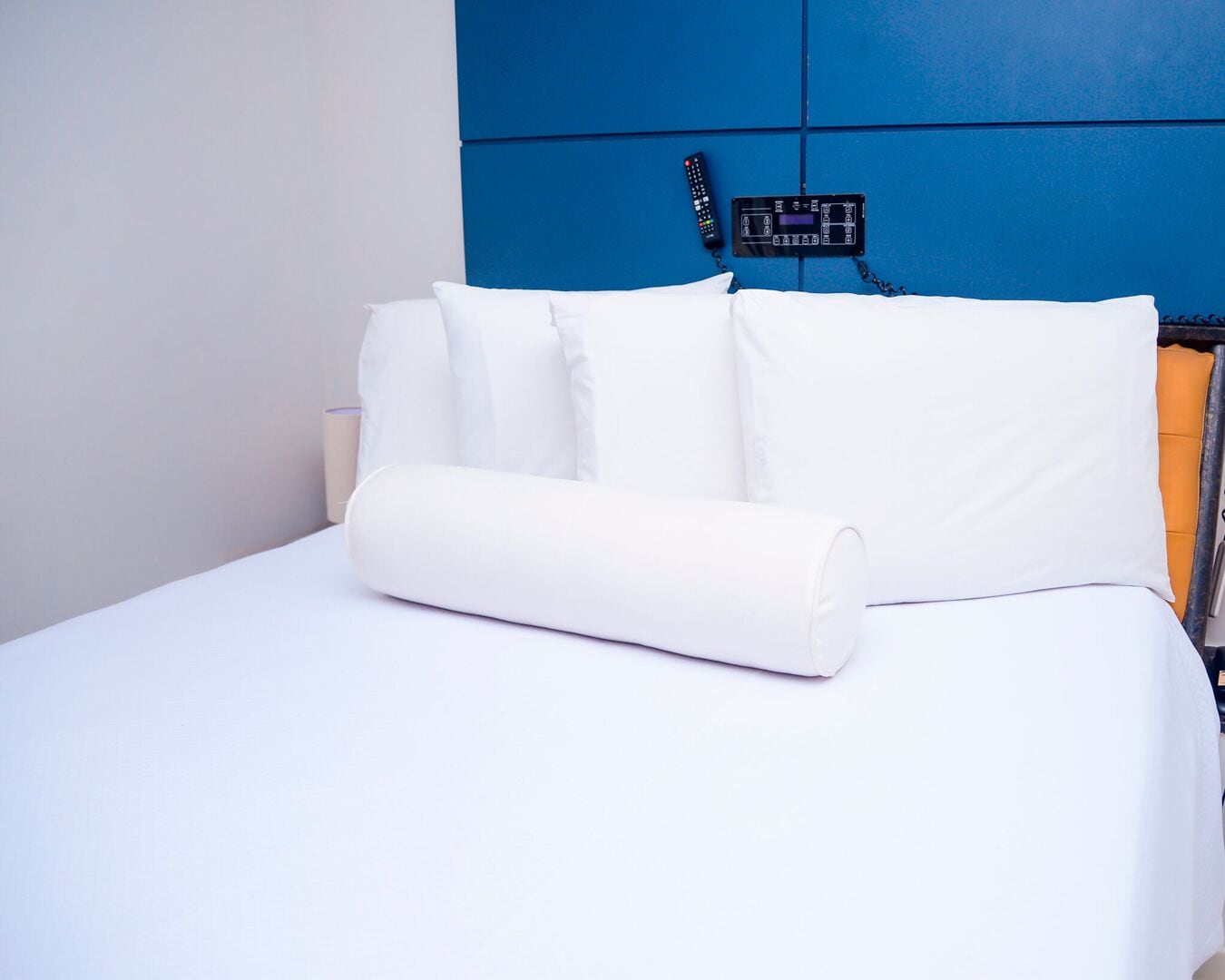 A neatly made bed with white linens and pillows, a remote control on the headboard, and a blue wall behind.