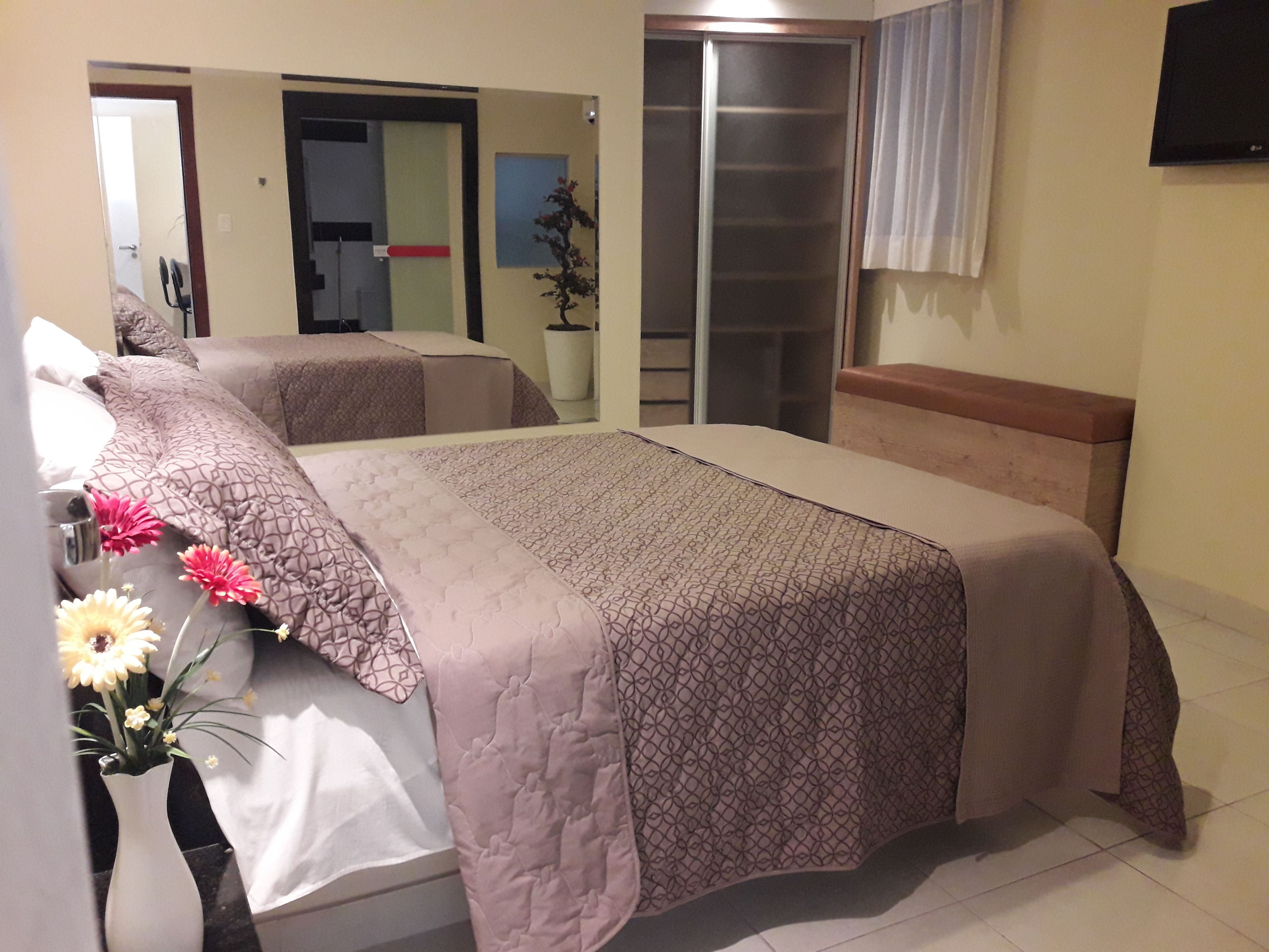 Un dormitorio con cama, mesita de noche y espejo. Hay una planta en maceta en el suelo y una ventana con cortinas.