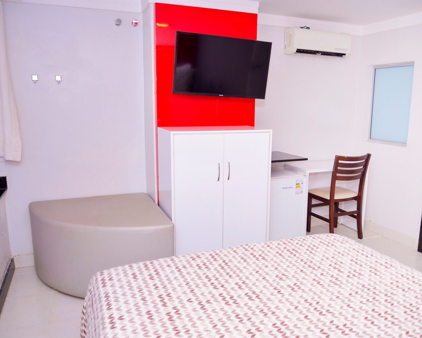 A bedroom with a bed, a desk with a chair, a wardrobe, a television, and an air conditioning unit.