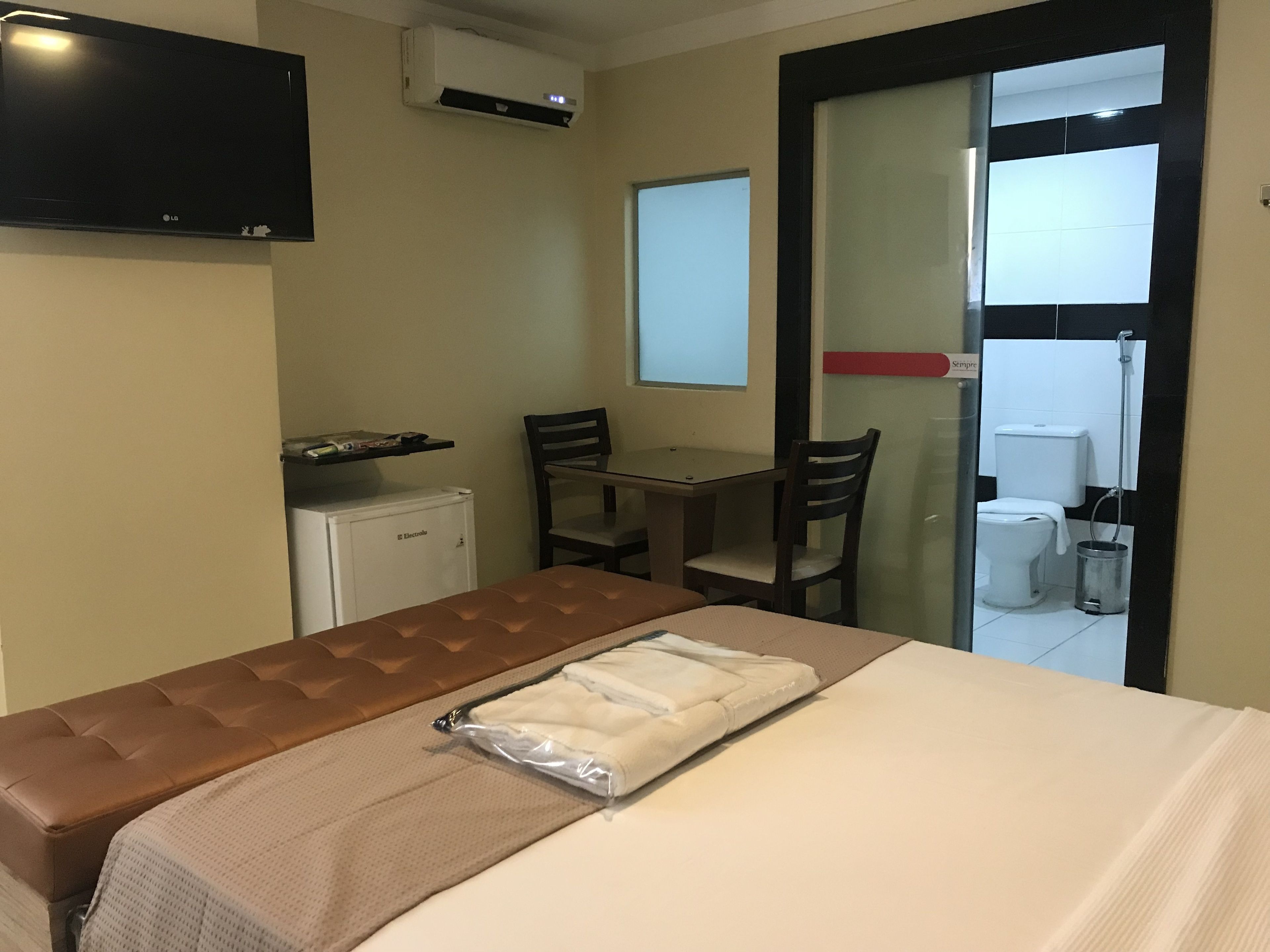 Ein Hotelzimmer mit Bett, kleinem Kühlschrank, Fernseher, Tisch mit Stühlen und einem durch eine Glastür einsehbaren Badezimmer.