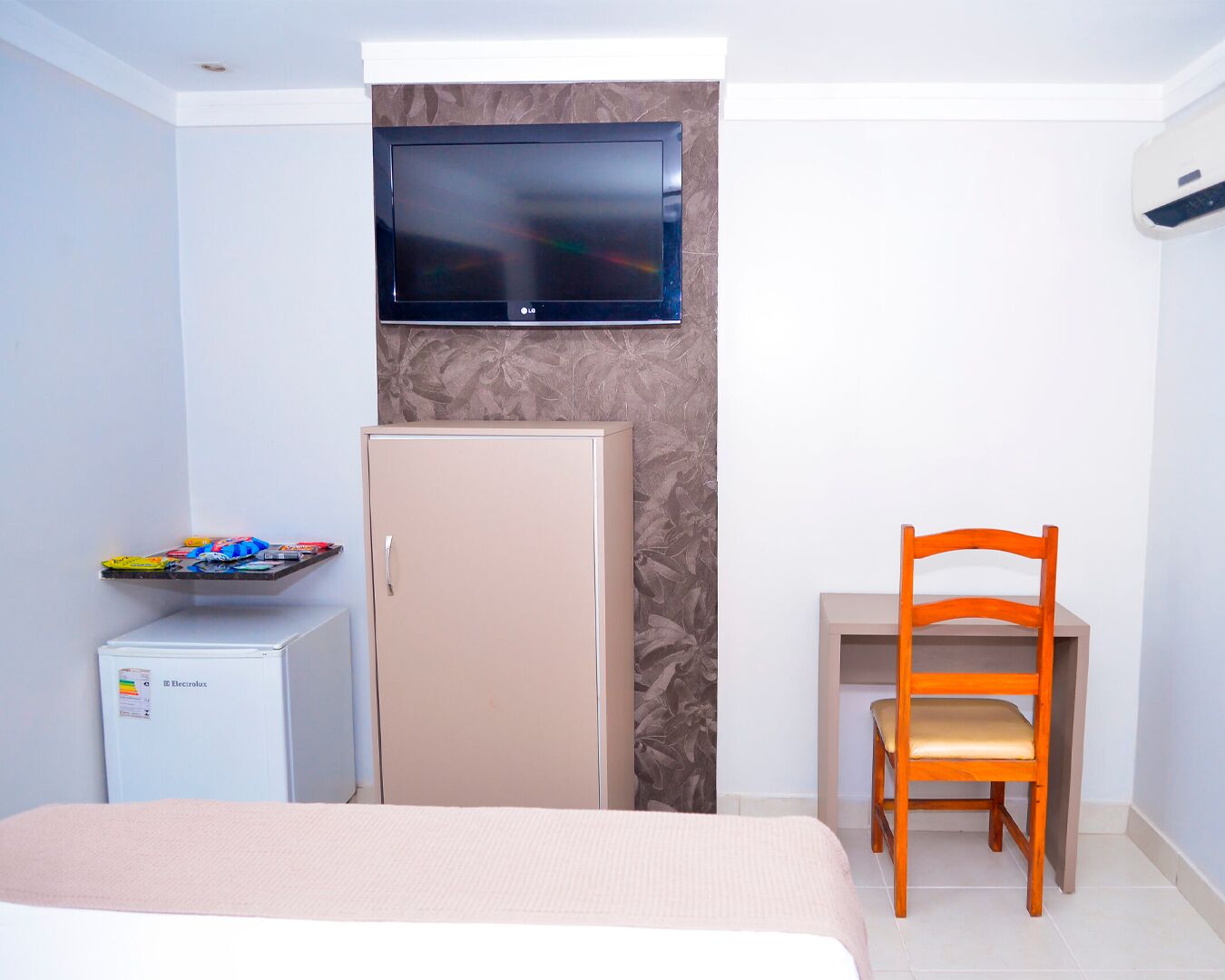 A hotel room with a bed, a desk with a chair, a television mounted on the wall, a minibar, and an air conditioning unit.
