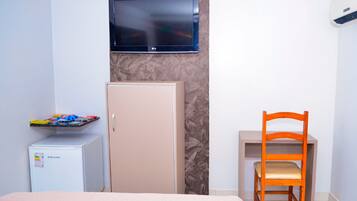 A hotel room with a bed, a desk with a chair, a television mounted on the wall, a minibar, and an air conditioning unit.