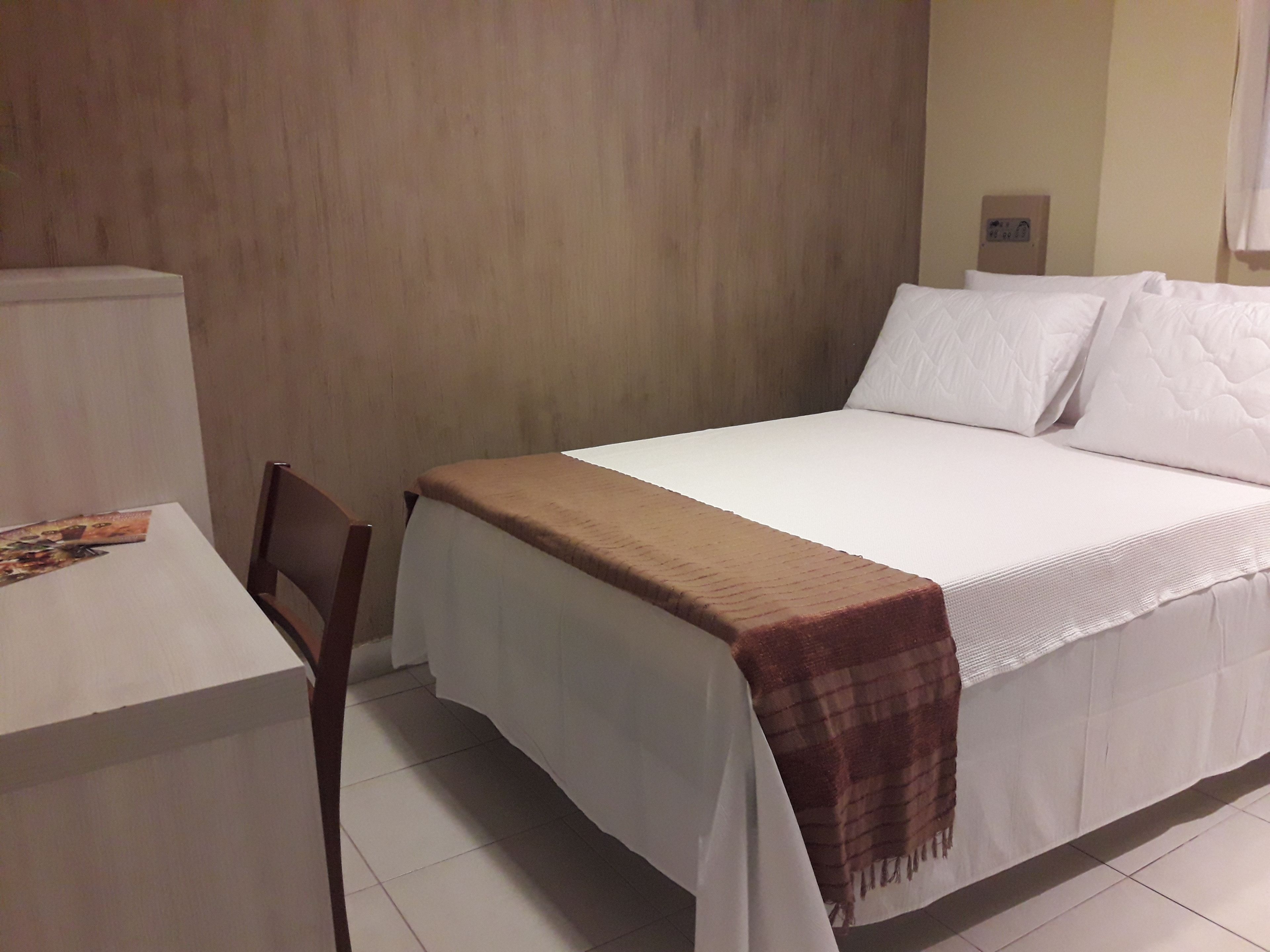 Una cama bien hecha con edredón blanco y almohadas, una manta marrón, una silla de madera y una mesita con una revista.