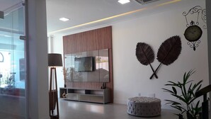 Ein modernes Wohnzimmer mit Flachbildfernseher, einer Wand aus Holzpaneelen, einem runden Ottomanen und Wandkunst mit Blättern.