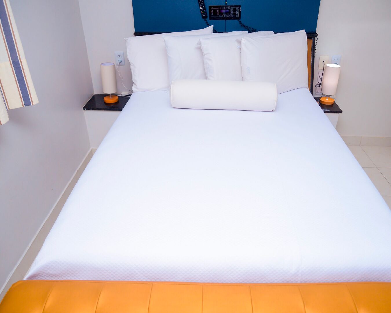 A neatly made bed with white bedding and an orange headboard. There are two bedside tables with lamps and a digital device on one of them.