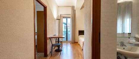 A narrow hallway with wooden flooring, a dining area with a table and chairs, a bed, and a bathroom with a sink and mirror.