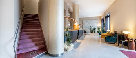 A modern hotel lobby with a marble staircase, a reception desk, seating area, and large columns.