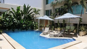 A swimming pool with lounge chairs and umbrellas, surrounded by greenery and a building with large windows. - The Blue Hotel. (Cartagena)