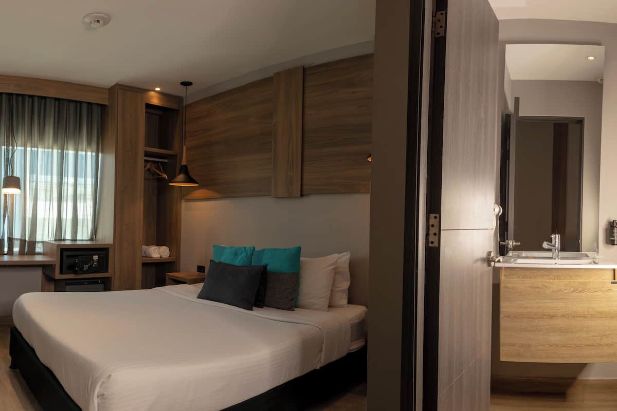 A modern hotel room with a large bed, a desk with a chair, a wardrobe, and a bathroom with a mirror and sink.