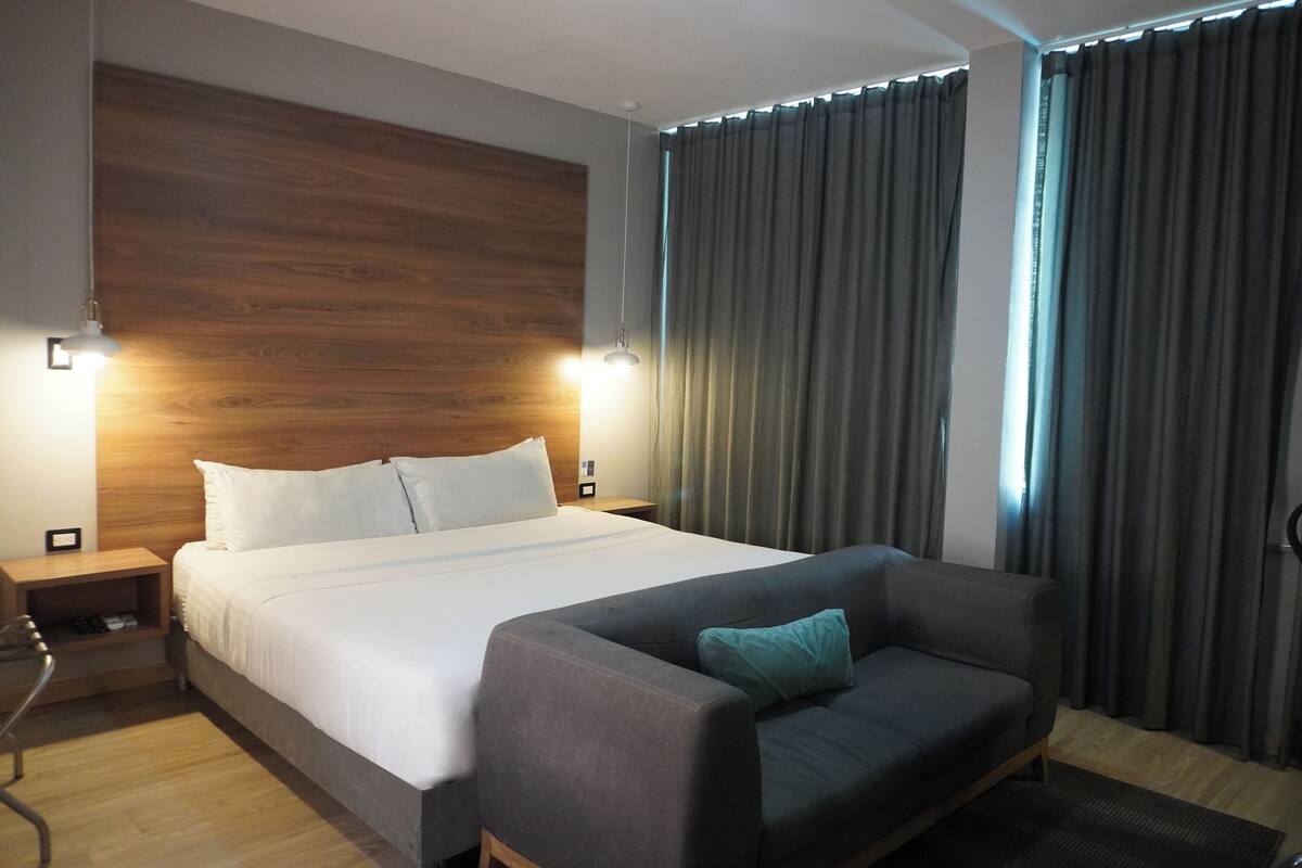 A modern hotel room with a large bed, a sofa, wooden headboard, and curtains.