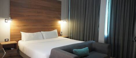 A modern hotel room with a large bed, a sofa, wooden headboard, and curtains.