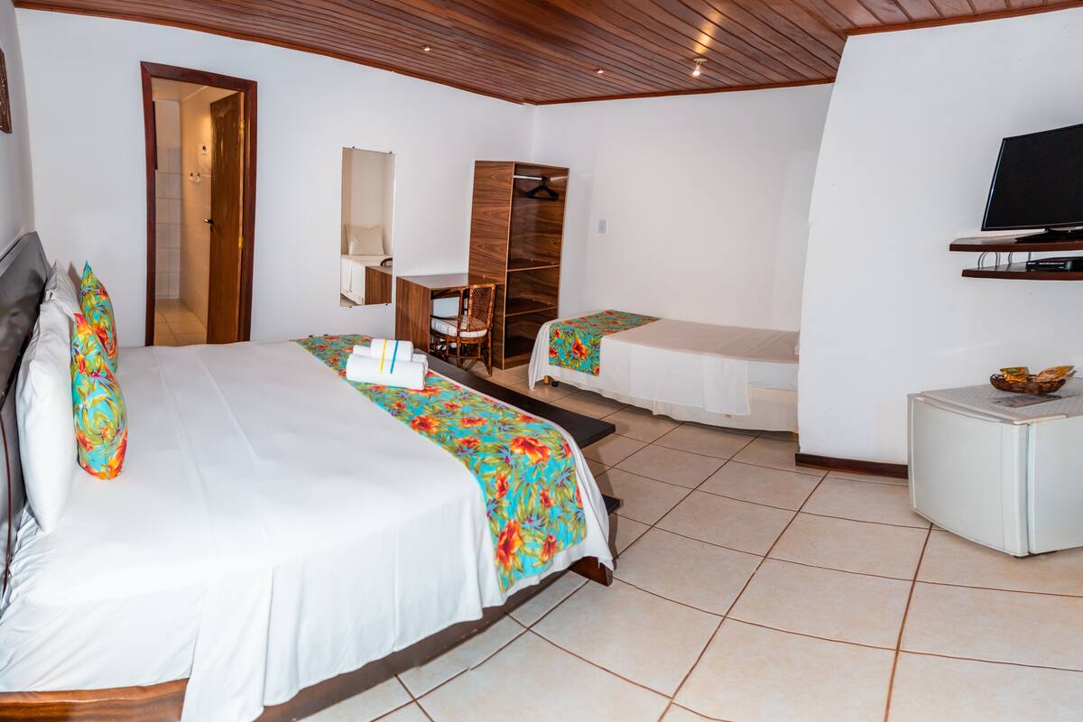 A hotel room with a large bed, a smaller bed, a desk, a chair, a TV, and a minibar.