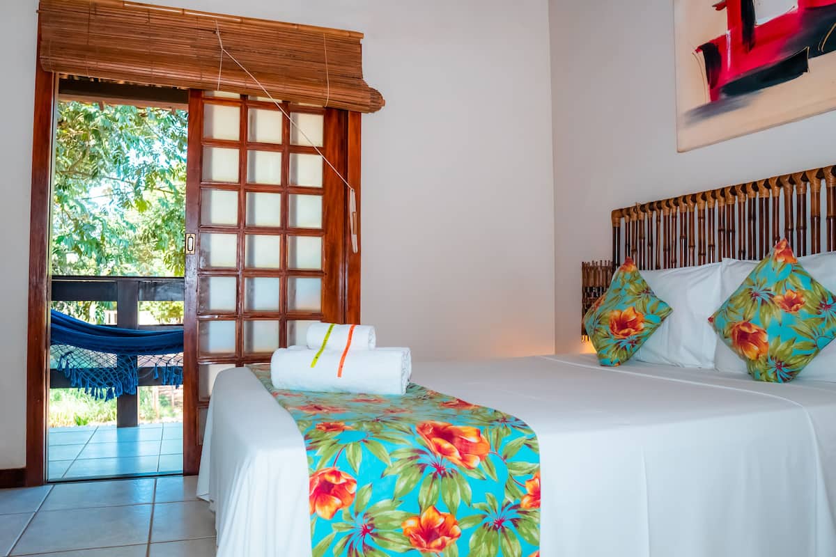 A hotel room with a bed, a hammock, a window with a view of greenery, and a painting on the wall.