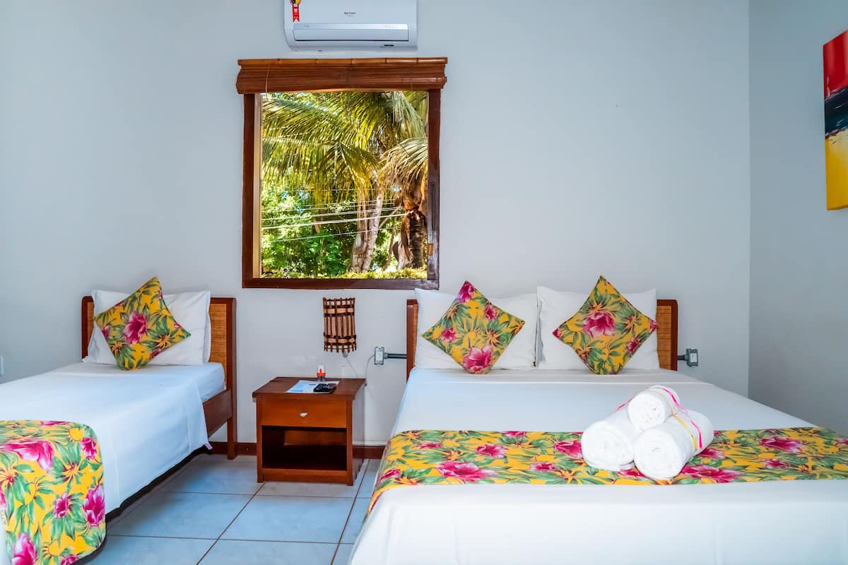 A hotel room with two beds, each with floral bedding, a window with a view of greenery, and an air conditioning unit above the window.