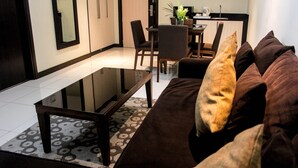 Een moderne woonkamer met een glazen salontafel, een bruine bank, een vloerkleed met patroon, en een eethoek met stoelen, met een kleine keuken op de achtergrond.